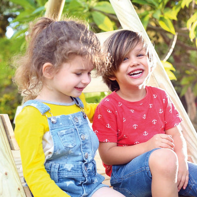 Two children enjoy a sunny day on the TP Toys Knightswood Double Wooden Swing & Slide Set. The girl in denim overalls and the boy in a red anchor-print shirt with denim shorts smile as they play outdoors.