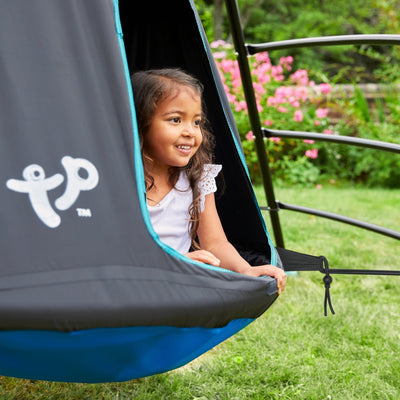 A young girl smiles as she sits inside the TP Toys TP UFO Den & Climbing Frame, surrounded by green grass and pink flowers, enjoying outdoor play in the garden.