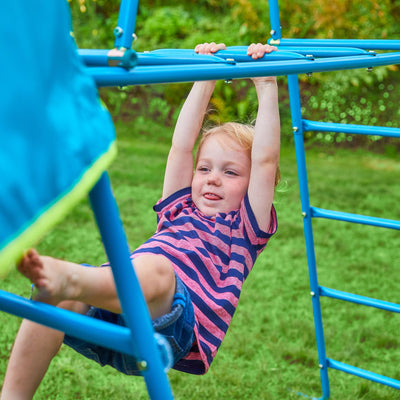 A young child in a striped shirt hangs from the blue TP Explorer Metal Climbing Frame Set & Jungle Run by TP Toys, surrounded by green grass and foliage outdoors.