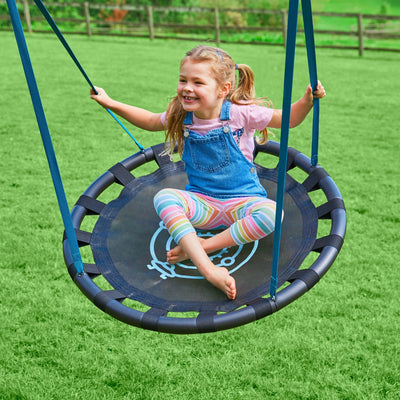 A young girl smiles while sitting on the nest swing of the TP Knightswood Double Wooden Swing Set with Basketball Hoop by TP Toys in a grassy outdoor area.