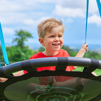 A young boy in a red shirt smiles on the TP Toys Knightswood Double Wooden Swing Set with Nest Swing & Basketball Hoop, holding blue ropes outdoors with greenery and a cloudy sky in the background.
