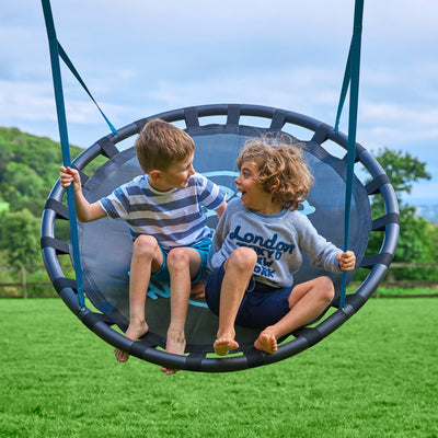 Two kids enjoy the TP Toys Deluxe Double Metal Swing featuring a giant nest seat, while a basketball hoop stands nearby for extra fun.
